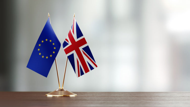 European Union And British Flag Pair On A Desk Over Defocused Background